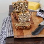 Nussbrot auf Schneidebrett mit Messer, Butter im Hintergrund, auf einem Holztisch mit Tischdecke.