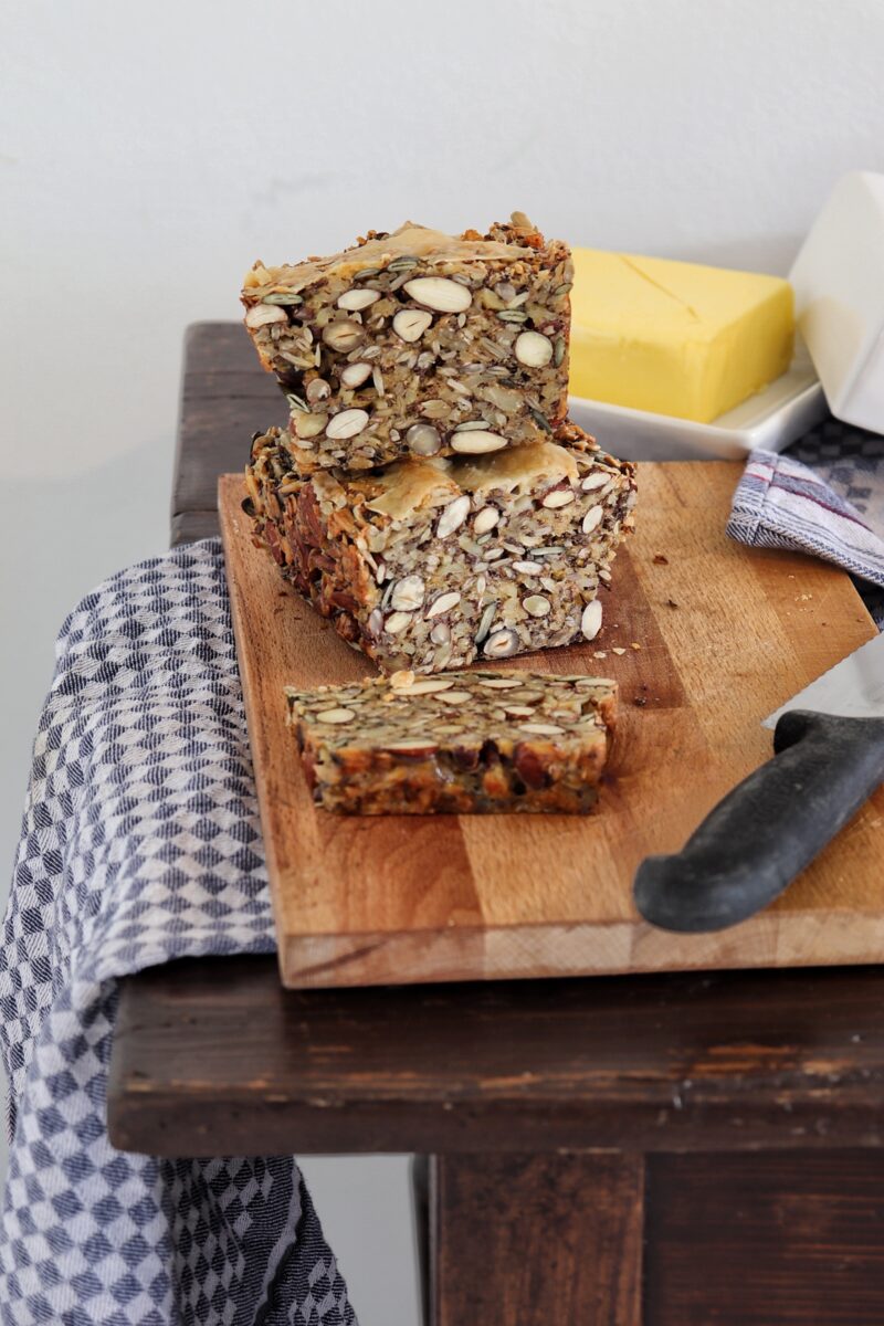 Nussbrot auf Schneidebrett mit Messer, Butter im Hintergrund, auf einem Holztisch mit Tischdecke.