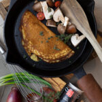 Pfannkuchen in einer Gusseisenpfanne mit Pilzen und Tomaten, Holzspatel daneben. Zwiebeln und Gewürz im Vordergrund.