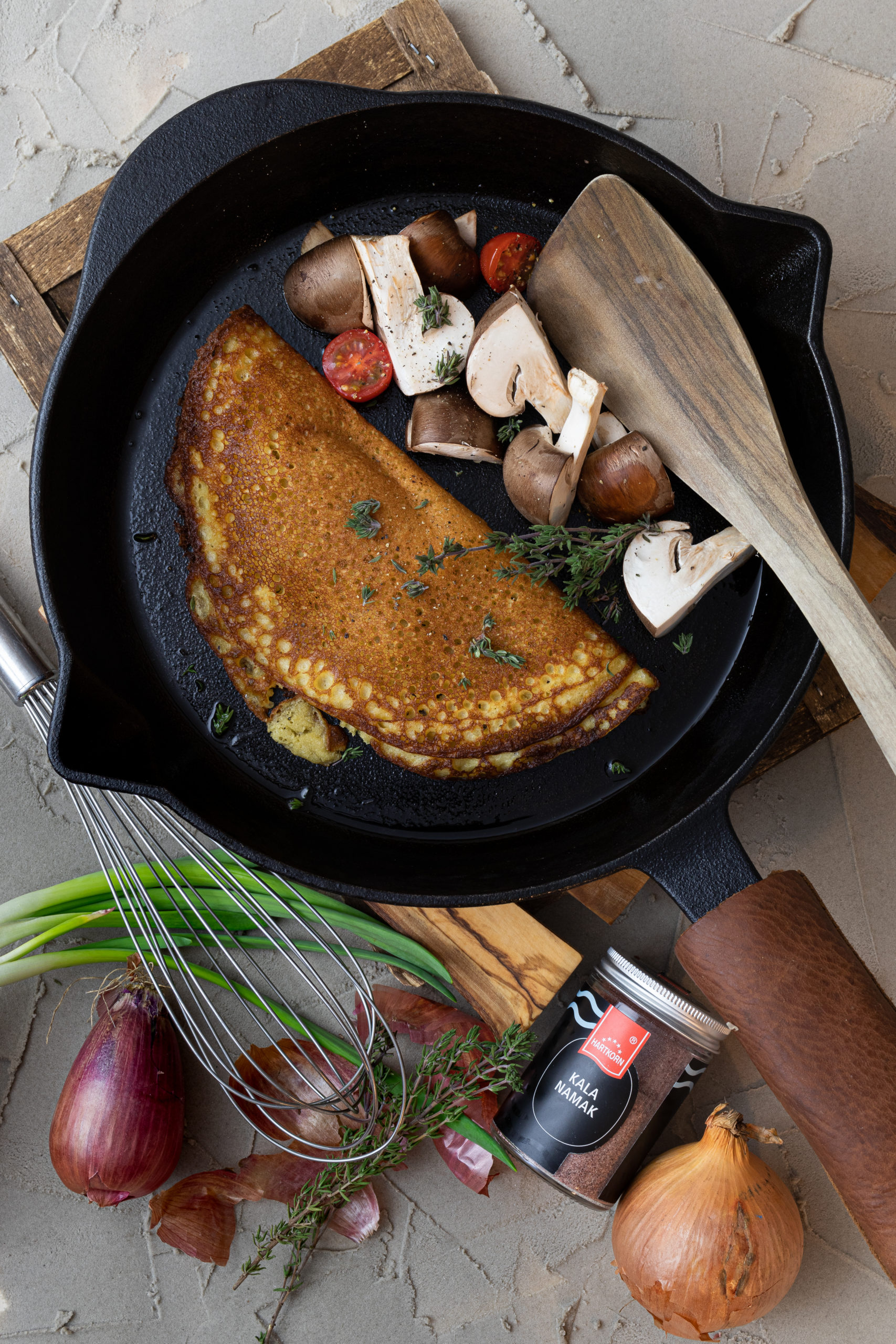 Pfannkuchen in einer Gusseisenpfanne mit Pilzen und Tomaten, Holzspatel daneben. Zwiebeln und Gewürz im Vordergrund.