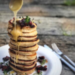 Ein Stapel Pfannkuchen mit Honig, garniert mit Kräutern und Speck, auf weißem Teller. Löffel gießt Honig darüber.