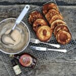 Pfannkuchen auf einem Rost neben Schüssel mit Teig, Gabel, Messer sowie Ahornsirup und Vanilleextrakt auf Holztisch.