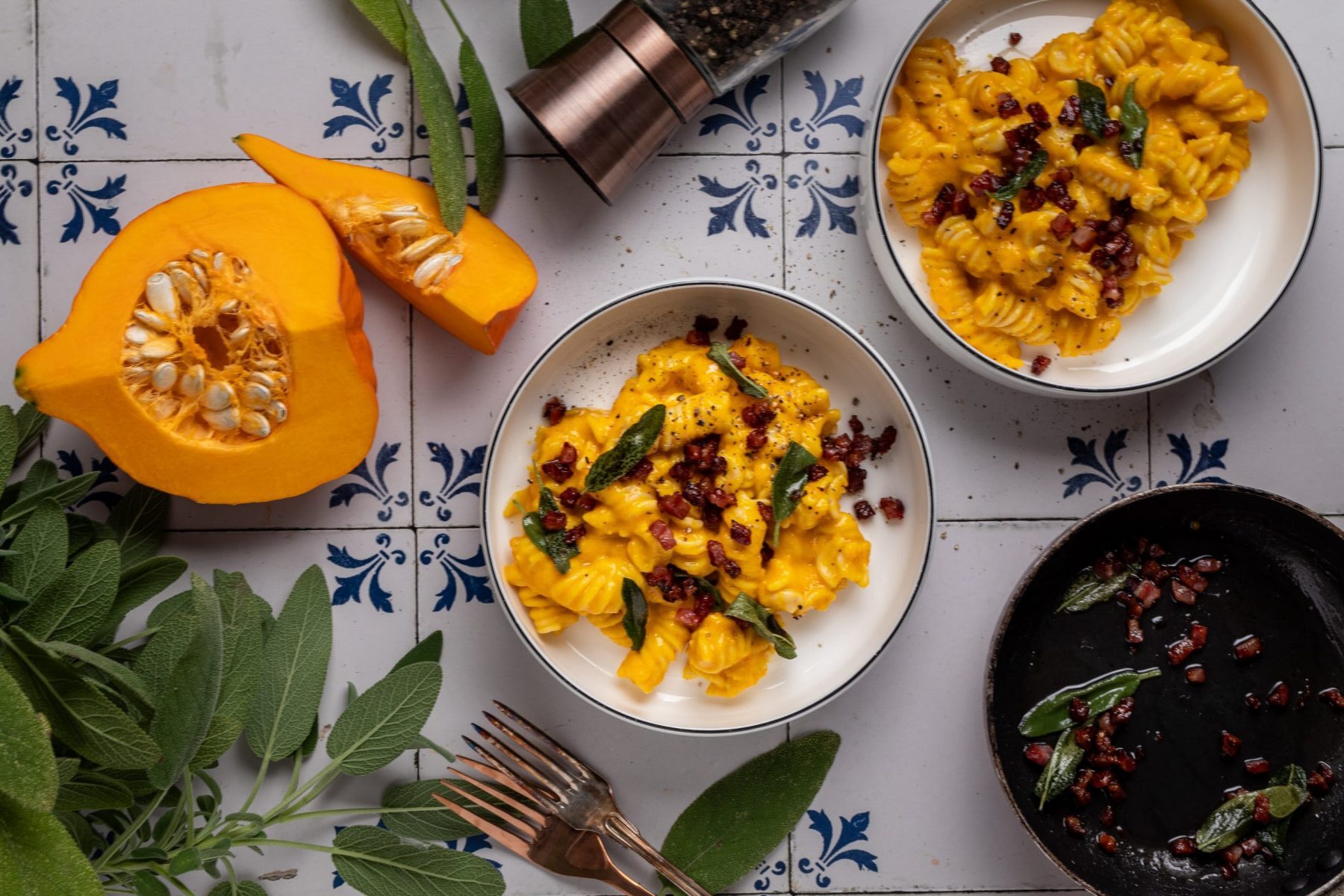 Kürbis-Pasta mit Speck und Salbei – Herbstlicher Genuss Nudeln mit Kürbissauce, Speck und Salbei in Schalen auf Fliesen, neben Kürbis, Pfefferstreuer und frischem Salbei.