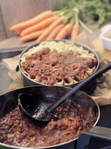 Nudelgericht mit Hackfleischsoße in Auflaufform, daneben dampfende Pfanne mit Soße, im Hintergrund Karotten.