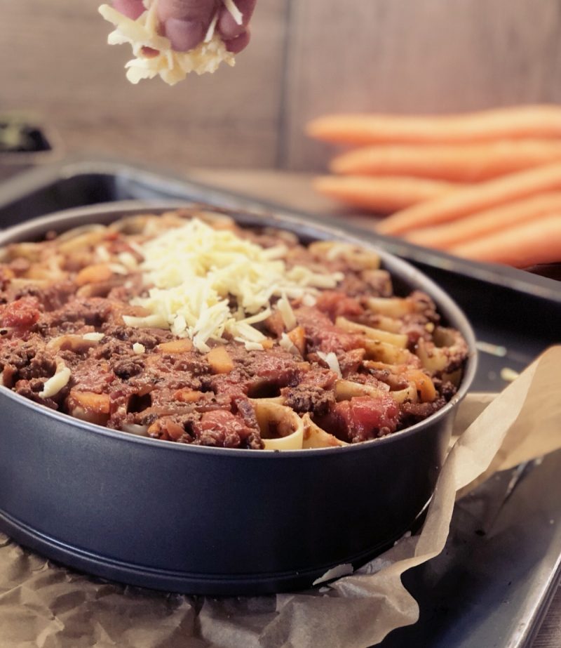 Ein runder Auflauf mit Nudeln, Hackfleisch und Tomaten in einer Backform, darüber wird geriebener Käse gestreut.