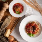 Teller mit Fleischbällchen auf Pasta und in Tomatensoße, Dekoration mit Basilikum, umgeben von Zwiebeln und Käse auf Holzbrett.