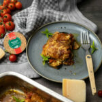 Ein Teller mit einer Portion überbackener Lasagne, daneben Tomaten, Parmesan, eine Gewürzdose und eine Auflaufform.