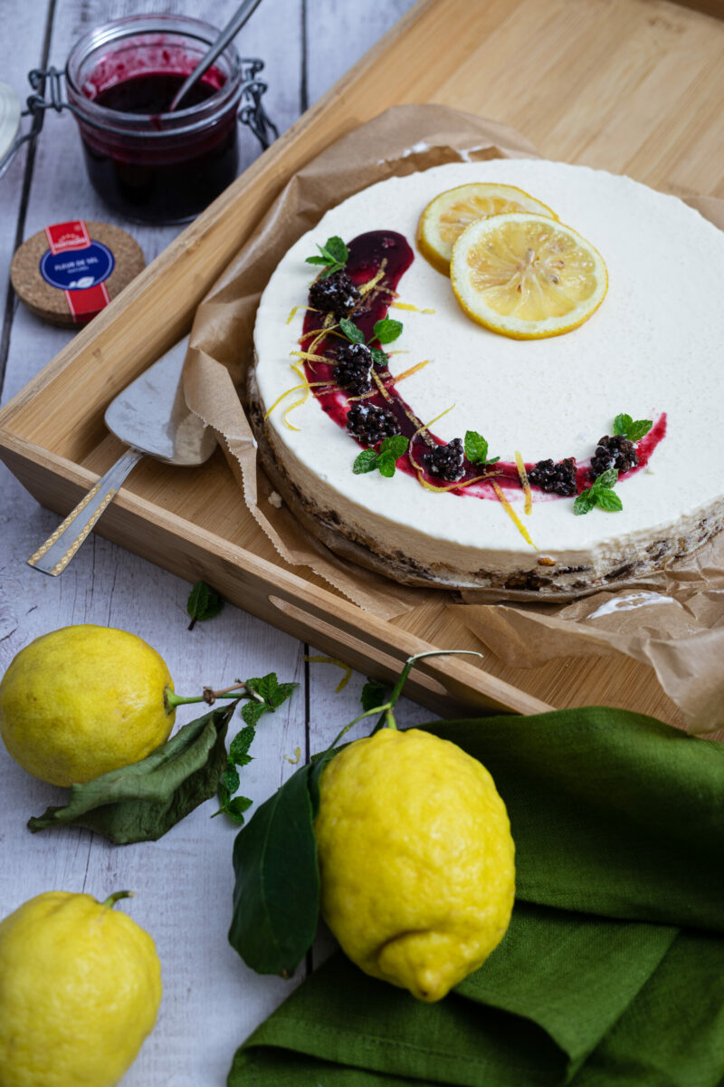 Zitronenkuchen dekoriert mit Zitronenscheiben, Beeren und Minze auf Holztablett, daneben ein Glas mit Beerenkonfitüre.