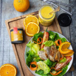 Salat mit Hähnchen, Orangen und Karotten auf einem Holzbrett. Daneben ein Glas Dressing und Curry-Gewürz.