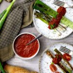 Zwei Teller mit gegrilltem Lauch, Tomatensauce und Tomaten, daneben eine Schüssel Sauce und Brot auf einem Tuch.