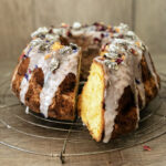 Ein Gugelhupf mit Zuckerguss und essbaren Blüten auf einem Gitter. Ein Stück wurde herausgeschnitten.