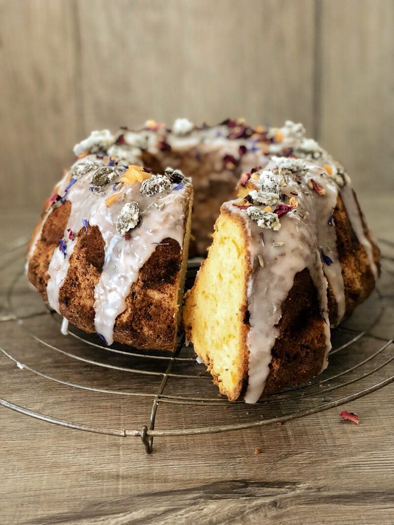 Ein Gugelhupf mit Zuckerguss und essbaren Blüten auf einem Gitter. Ein Stück wurde herausgeschnitten.