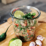 Ein Glas mit frischem Salsa-Dip, garniert mit Limettenscheiben und Kräutern, daneben Limette, Knoblauch und Chili auf Holzbrett.