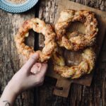 Hand greift nach einem gebackenen Kringel mit Sesam auf einem Holzbrett, daneben ein Teller mit Sesamkörnern.