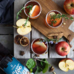 Drei Cocktails auf einem Holzbrett mit Äpfeln und Rosmarin garniert, umgeben von Gewürzen und einer Gin-Flasche.