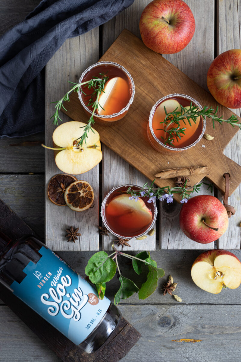 Drei Cocktails auf einem Holzbrett mit Äpfeln und Rosmarin garniert, umgeben von Gewürzen und einer Gin-Flasche.
