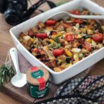 Ofenfrisches Gemüsegericht mit Tomaten, Paprika und Kräutern in weißer Form, daneben Kamera und Gewürzdose auf Holztisch.