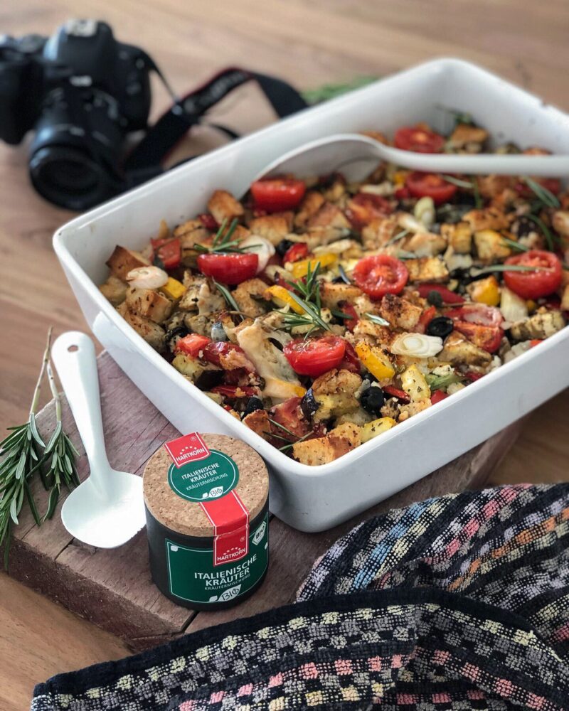 Ofenfrisches Gemüsegericht mit Tomaten, Paprika und Kräutern in weißer Form, daneben Kamera und Gewürzdose auf Holztisch.