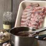 Topf mit dunkler Sauce und Schneebesen, daneben Knoblauch und rohe Rippchen in einer weißen Schale auf Holztisch.