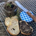 Ein Glas Olivenpaste mit Thymian, daneben Brotscheiben, eine bestrichen, ein Messer und ein blau-weiß kariertes Deckel.