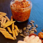 Glas mit rotem Pesto, daneben Penne-Nudeln, Kürbiskerne und eine Hand, die getrocknete Tomaten greift, auf schwarzem Untergrund.