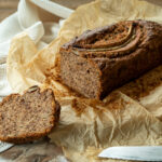 Bananenbrot auf Backpapier, mit einem abgeschnittenen Stück und einem Messer daneben auf einem Holztisch.