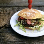 Burger mit Gemüse und Rucola auf einem Teller, dekoriert mit aufgespießten Tomaten, auf rustikalem Holztisch.