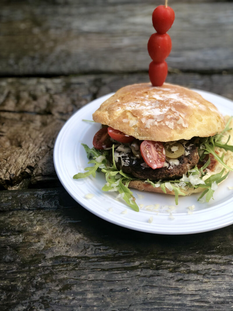 Burger mit Gemüse und Rucola auf einem Teller, dekoriert mit aufgespießten Tomaten, auf rustikalem Holztisch.