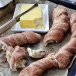 Frisch gebackene, gedrehte Brote auf Backpapier neben einem Teller mit Butter und Messer.