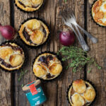 Herzhafte Mini-Tartes mit Ziegenkäse auf rustikalem Holzbrett, umgeben von roten Zwiebeln und Thymian.