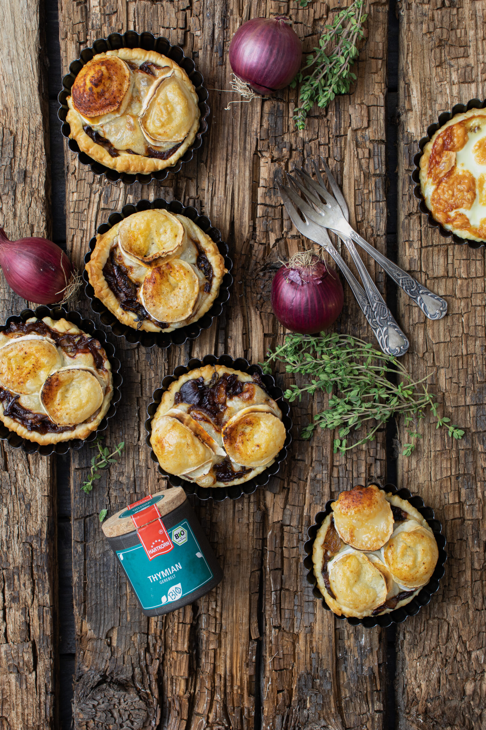 Herzhafte Mini-Tartes mit Ziegenkäse auf rustikalem Holzbrett, umgeben von roten Zwiebeln und Thymian.