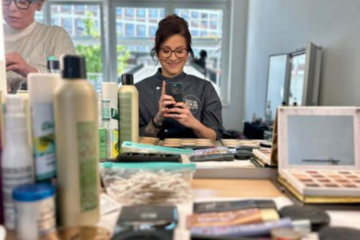 Frau in Brille macht Selfie vor Spiegel, umgeben von Kosmetikprodukten und Friseurzutaten in einem Salon.