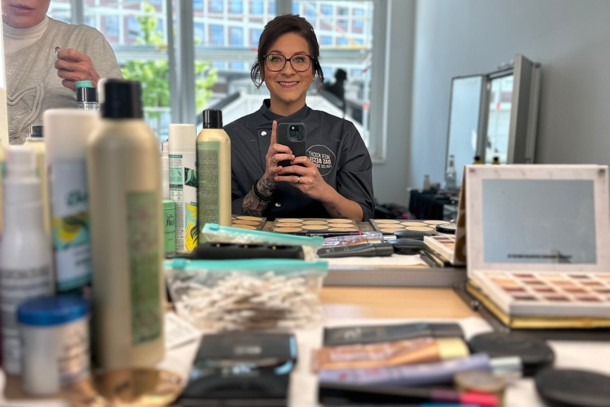 Kochshow-Sat.1-5 Person in einem Salon macht ein Selfie im Spiegel, umgeben von Pflege- und Kosmetikprodukten auf einem Tisch.