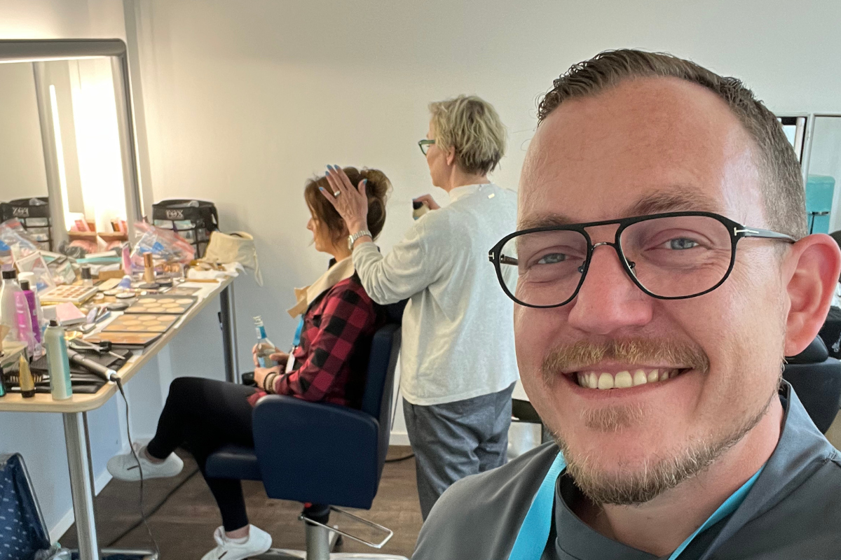 Kochshow-Sät.1-6 Ein Mann lächelt in die Kamera, während eine Frau im Hintergrund in einem Schminkraum gestylt wird.