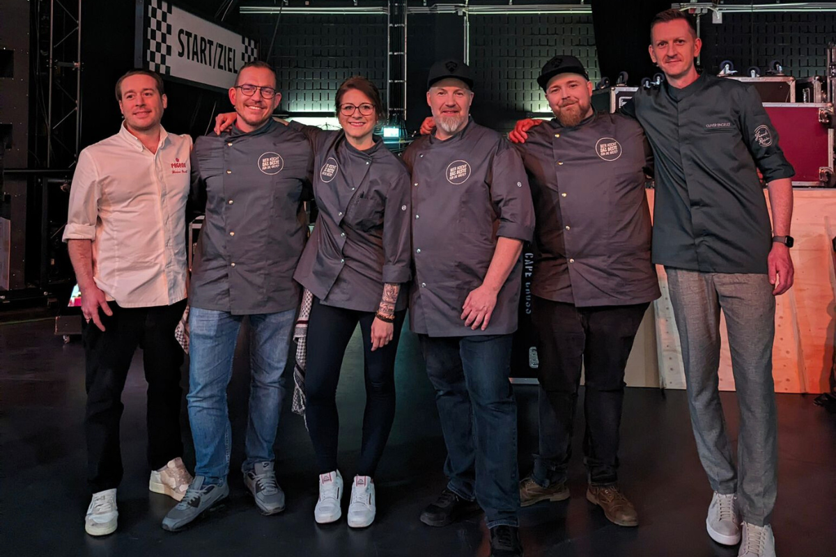 Kochshow-Sat.1-9 Sechs Menschen in Kochjacken posieren lächelnd zusammen auf einer Bühne vor einem