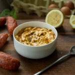 Schale mit cremiger, orangefarbener Sauce, umgeben von Wurst, Zitronen und Eiern auf einem Holzbrett. Ein Löffel liegt daneben.