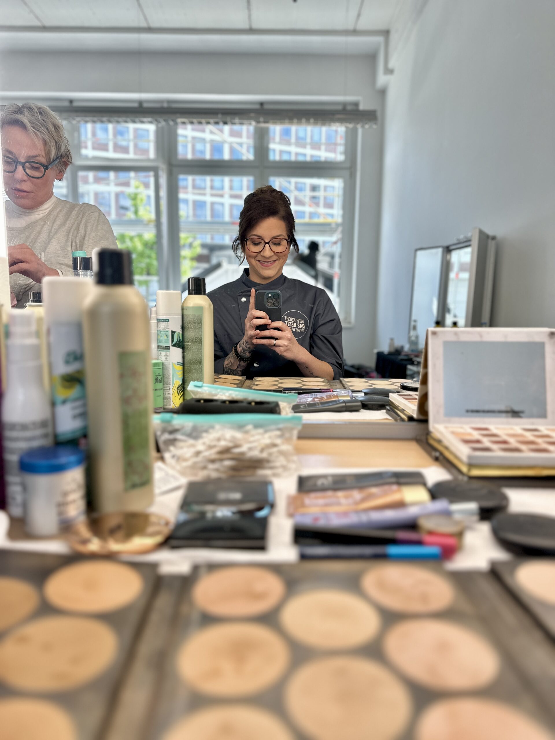 Zwei Personen im Friseursalon, umgeben von Kosmetikprodukten; eine Person macht ein Selfie im Spiegel.