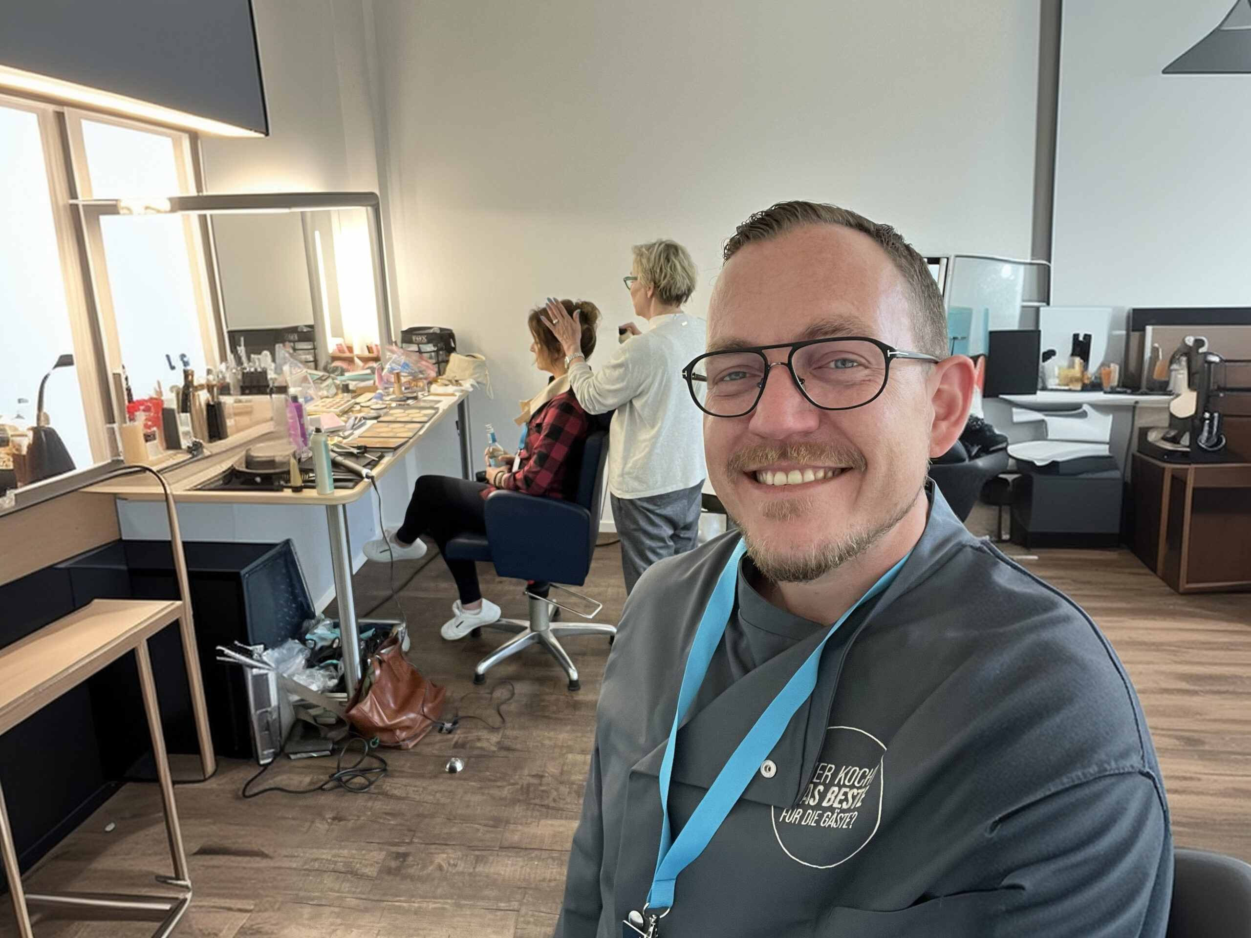 Mann lächelt in die Kamera im Friseursalon, während im Hintergrund eine Person frisiert wird.