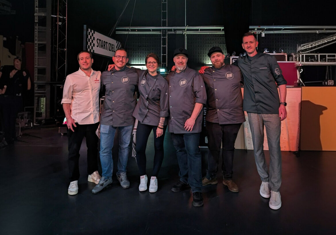 Sechs Personen posieren in Kochjacken auf einer Bühne vor einem