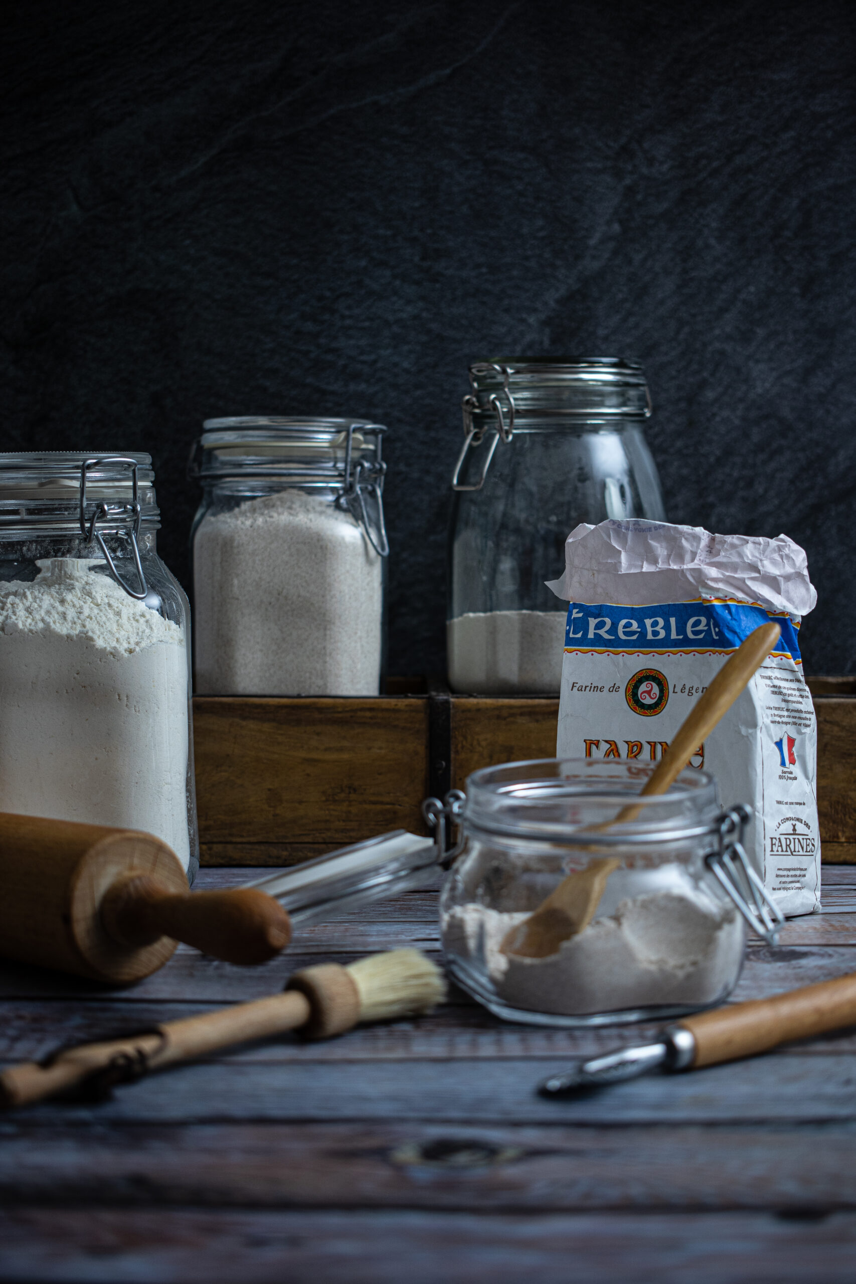 Verschiedene Vorratsgläser mit Mehl und Küchenutensilien wie Nudelholz und Pinsel auf einem Holztisch.