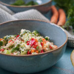 Schüssel mit frischem Couscous-Salat, gemischt mit Paprika, Gurken und Kräutern. Im Hintergrund Möhren und Stoff.