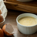 Eierkarton, zerbrochene Eierschalen und eine kleine Schüssel Creme auf Holztisch, im Hintergrund eine Suppe.