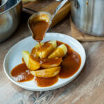 Gekochte Kartoffeln auf einem Teller werden mit brauner Soße aus einer Kelle übergossen. Im Hintergrund Töpfe.