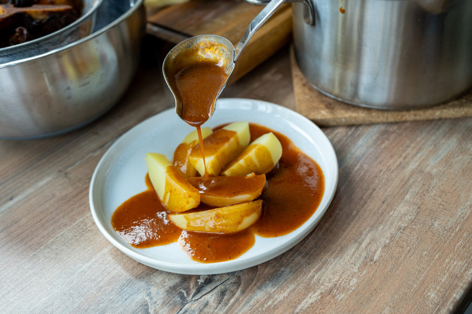 Gekochte Kartoffeln auf einem Teller werden mit brauner Soße aus einer Kelle übergossen. Im Hintergrund Töpfe.