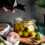 Glas mit Rosenkohl wird mit Öl übergossen. Holzbrett mit Lorbeerblatt neben Glas. Pflanze im Hintergrund.