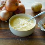 Schüssel mit gelber Suppe, Löffel, daneben Zwiebeln, Kartoffeln und gewellte Pommes frites auf Holzbrett.