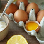 Eierkarton mit sechs braunen Eiern, eines geöffnet, daneben eine Schüssel mit Sahne und ein Zitronenstück.