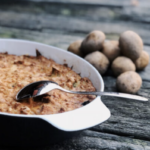 Auflaufform mit Kartoffelgratin und Löffel auf einem Holztisch, daneben liegen mehrere frische Kartoffeln.