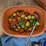 In einem Terrakotta-Topf sind gebackene Rosenkohl und Kirschtomaten mit frischen Kräutern und einem Löffel serviert.