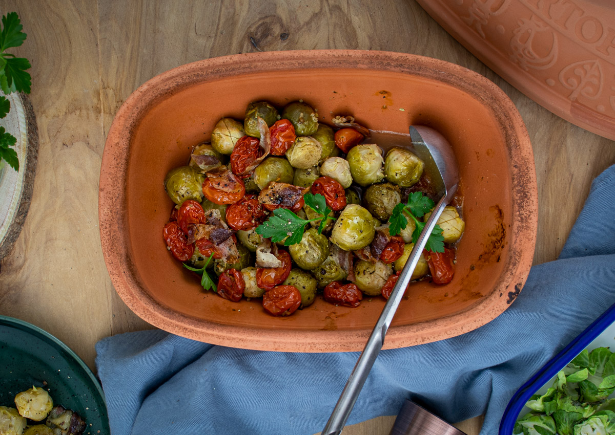 Rosenkohl_aus_dem_Römertopf_1 Gebratener Rosenkohl und Kirschtomaten in einer Terrakotta-Schale auf einem Holzbrett mit einem Löffel daneben.
